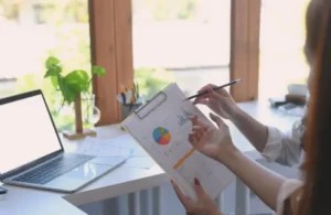 Two people discussing a clipboard with colorful charts near a laptop. Bright office setting with plants and natural light, conveying a focused business tone.