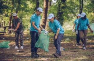 A group of volunteers in blue shirts and caps clean a forest, collecting litter into green bags. The scene conveys teamwork and environmental care.