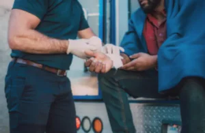 A person in a polo shirt and gloves is bandaging another person's hand. The seated person wears a red shirt and blue blanket, suggesting care and assistance.