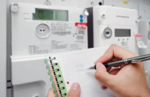A person writes numbers in a notepad while observing multiple electrical meters in the background. The scene conveys focus and record-keeping.