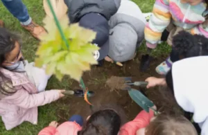 Children gathered around a small hole in the ground, joyfully planting a young tree. They hold shovels, surrounded by green grass.