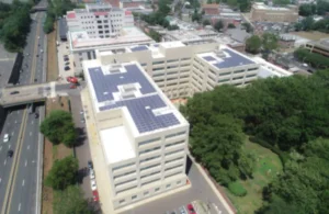 Aerial view of a large multi-story building with solar panels on the roof, surrounded by trees and roads. Nearby, cars are parked alongside the building.