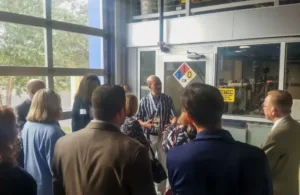 A group of people attentively listens to a presenter in a lab wearing a striped shirt. Warning signs on the door suggest a controlled environment.