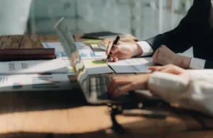 Two people work at a wooden table, focusing on charts and graphs. One writes notes, while the other uses a laptop, indicating teamwork and analysis.
