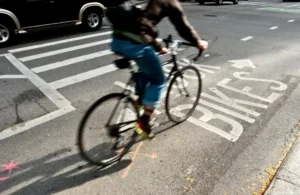 Cyclist in motion on a city street, riding through a marked bike lane. Blurred motion conveys speed and urban energy, with cars and crosswalk nearby.